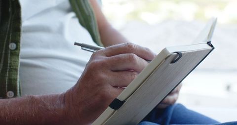 Mature man holding notebook and writing with metal pen, journaling by window light