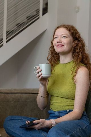 Red-haired woman relaxing with coffee and smartphone on couch at home, serene interior
