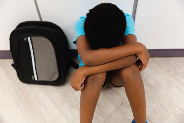 High angle view of sad african american elementary schoolboy sitting on floor in corridor. unaltered, childhood, education, sadness, loneliness, failure and school concept.