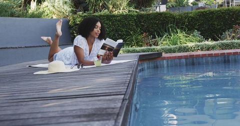 African woman relaxing and reading paperback on sunny pool deck with hat and drink
