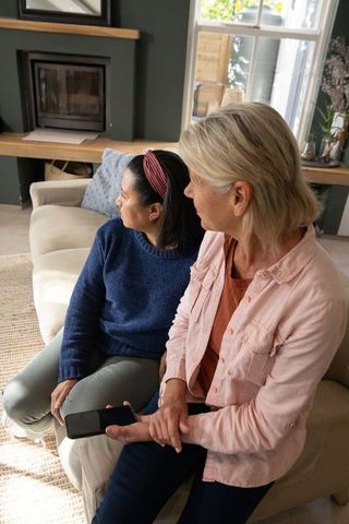 Multicultural women sitting together on cozy sofa holding smartphone sharing quiet moment