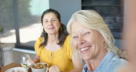 Smiling senior woman sharing lunch and wine with friend at home kitchen table