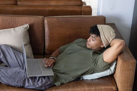 African american man relaxing on leather sofa using laptop for remote work and online learning