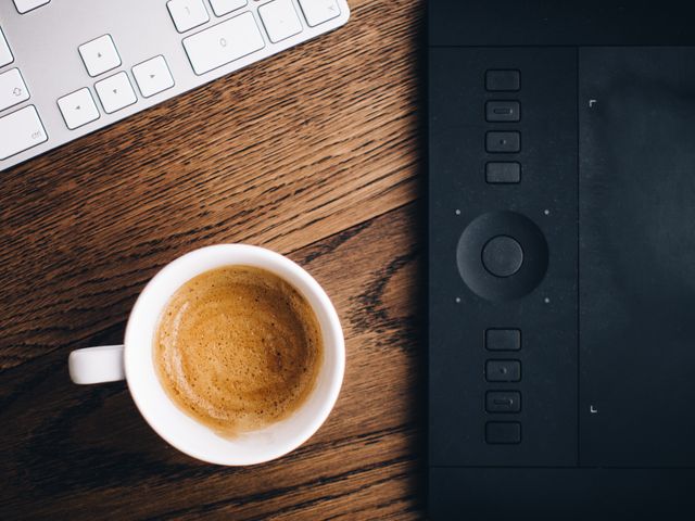 Coffee cup on a wooden desk next to a keyboard and drawing tablet. Useful for illustrating creative workspaces, freelancers, productivity. Ideal for blogs about coffee culture, home office setups, or articles on balancing work and breaks.