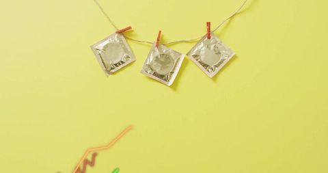 Hanging silver foil condom packets on twine with red clips over pale yellow background