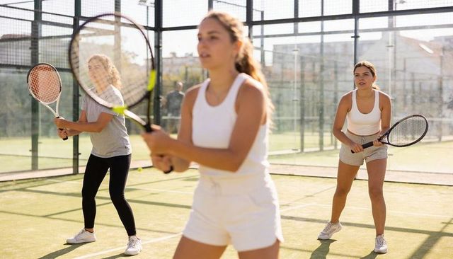 Three women playing tennis on sunlit glass court, urban team training and fitness