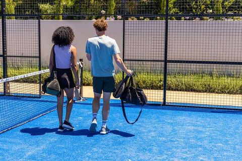 Young couple walking on blue padel court carrying rackets and sports bags in sunlight