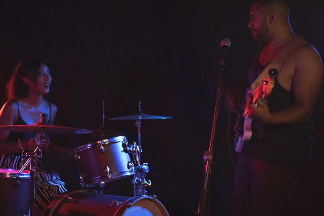 Man playing bass guitar and singing into microphone while female drummer accompanies him on stage in nightclub. Ideal for use in articles about live music, nightlife, entertainment venues, and band performances.