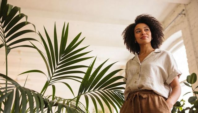 Confident woman standing among indoor palms in bright loft, relaxed minimal home workspace
