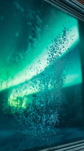 Vertical video revealing frosted window pane with glowing northern lights beyond night sky