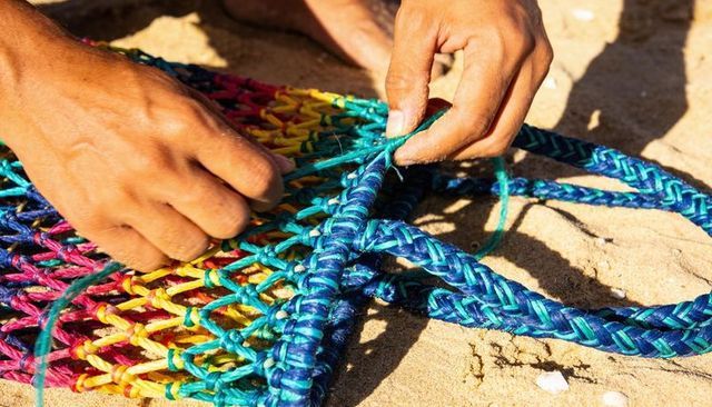 Hands weaving colorful fishing net on sandy beach, closeup mending braided blue rope mesh