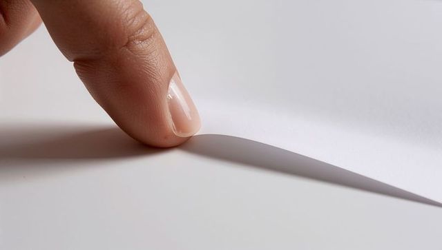 Fingertip lifting curled corner of white paper casting soft shadow, minimal macro detail