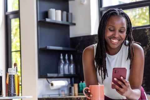 African woman leaning on marble kitchen counter holding smartphone and coffee mug, smiling