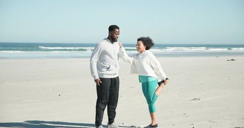 Black couple stretching and laughing on sunny beach shore, woman balancing leg in activewear