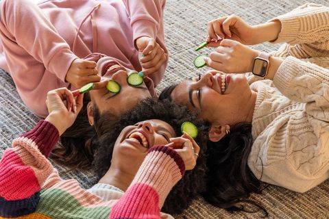 Diverse women lying on rug holding cucumber slices laughing during cozy home spa night