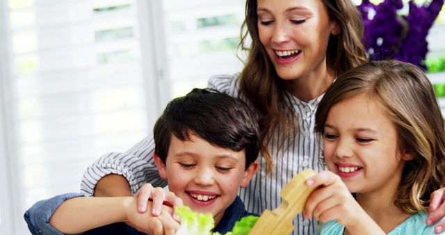 Cheerful mother bonding with her children while preparing a salad together in a modern home. Ideal for use in advertisements promoting family values, healthy eating habits, parenting tips, and lifestyle blogs. Displays themes of togetherness, joy, and a wholesome lifestyle.
