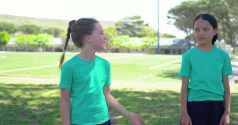 Two young girls chatting on sunny school sports field wearing green t-shirts
