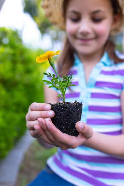 Vertical of smiling caucasian girl holding flower to plant in garden, selective focus, copy space. Health, childhood, ecology and happiness concept.