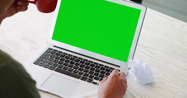 Person using a laptop with a green screen, suitable for inserting custom content for visual designs, advertisements, or presentations. Coffee mug and crumpled paper suggest a casual work environment, ideal for themes around remote work, studying, or tech usage.