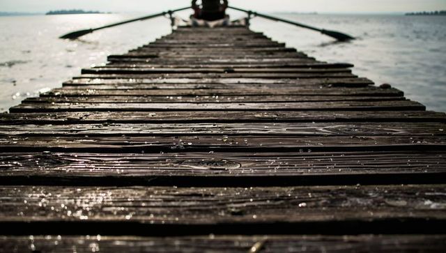 Weathered wooden pier leading to rowboat on lake with silhouetted oars, sparkling water