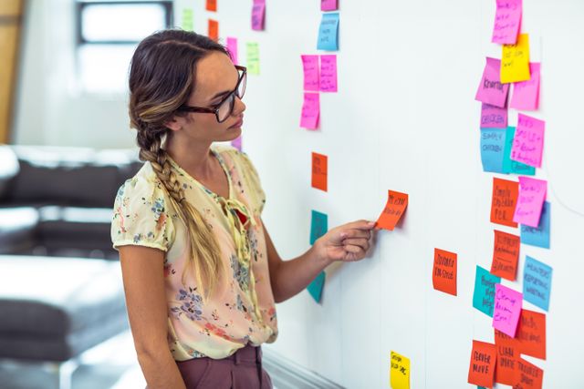 Woman organizing colorful sticky notes on whiteboard in office. Ideal for illustrating concepts of planning, brainstorming, project management, and teamwork in a professional setting. Useful for business presentations, articles on productivity, and workplace strategy visuals.