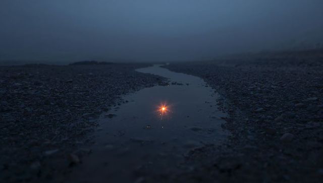 Warming amber reflection in narrow pebbled shore puddle guiding eye toward misty horizon