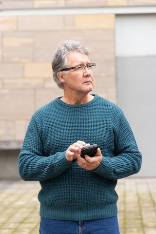 Thoughtful senior man using smartphone in urban plaza wearing teal sweater and glasses