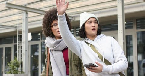 Friends hailing ride outside modern glass building while holding smartphone and bags