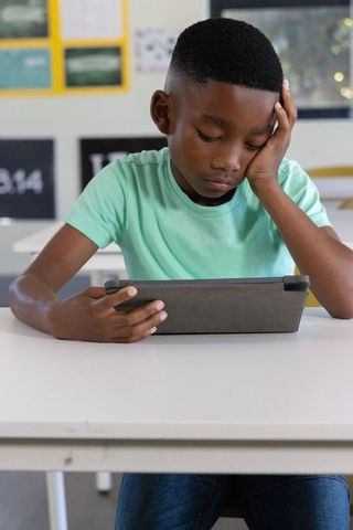 School-age african american boy using tablet at desk looking bored digital learning