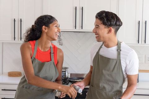 African american couple cooking and laughing together in modern bright kitchen with aprons