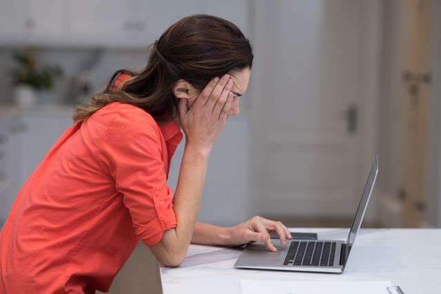 Woman in casual wear working on laptop at home office desk, appearing stressed and anxious. Ideal for use in articles about remote work challenges, mental health, work-life balance, and productivity tips.