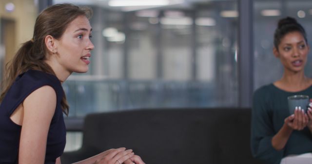 Two women are engaged in a serious conversation in a modern office environment. One woman appears to be intently listening while expressing her point, while the other holds a cup, participating thoughtfully. This image can be used in articles and websites focusing on business communication, workplace dynamics, corporate meetings, teamwork, and professional collaboration.