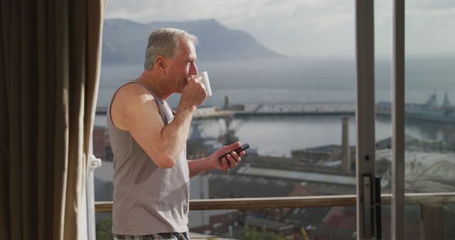 Caucasian senior man drinking coffee and using smartphone on sunny balcony. Retirement, senior lifestyle, happiness, domestic life, communication and wine making, unaltered.
