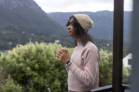 Nonbinary person standing on balcony holding mug, gazing at mountain valley, contemplative morning