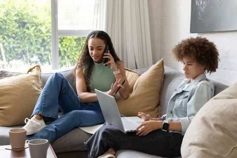 Two african american women chatting on phone and working on laptop on cozy sofa