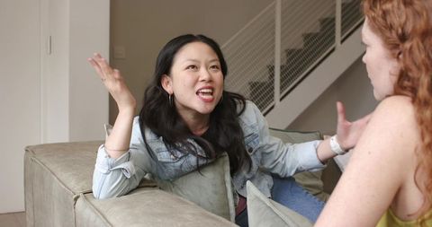 Two friends having engaging conversation on sofa, asian woman leaning and gesturing
