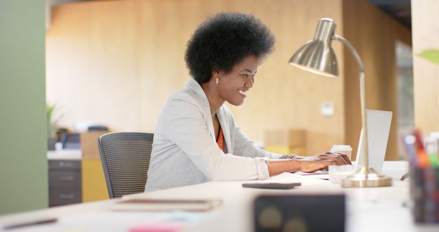 Professional woman with afro hair working on laptop in modern office. Ideal for advertisements or articles relating to workplace productivity, diversity in the workplace, business settings, and office-related content. Great for promoting office furniture, corporate environments, and tech usage in professional settings.