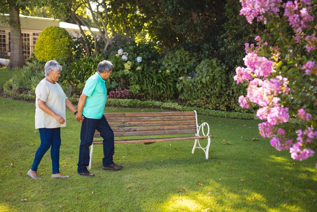 This image depicts a senior couple holding hands and walking in a park, showcasing love and togetherness in their retirement years. Ideal for use in advertisements or articles related to senior living, retirement communities, healthy aging, and outdoor activities for the elderly. The serene park setting with blooming flowers and lush greenery adds a peaceful and joyful ambiance.