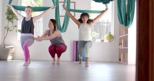 Three women practicing aerial yoga in a brightly lit studio, guided by an instructor. They are holding onto aerial silks while performing balancing poses. One woman is receiving hands-on assistance from the instructor. This image can be used to promote fitness classes, yoga studios, healthy lifestyle content, exercise programs, or wellness retreats.