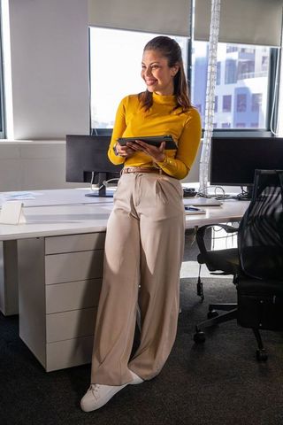 Confident businesswoman leaning on desk holding tablet and smiling in bright modern office
