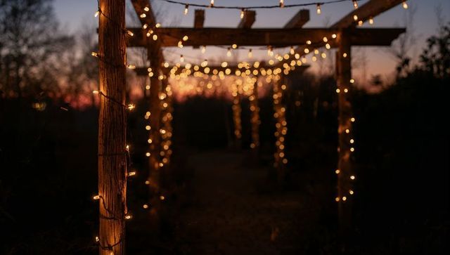 Glowing string lights wrapping wooden pergola posts at dusk creating warm bokeh path