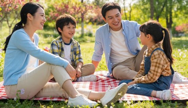 Family enjoying picnic in sunny park on red-and-white blanket, smiling and talking