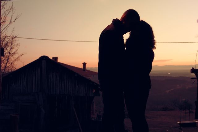 Silhouette of couple sharing an intimate embrace during sunset in a countryside setting. Ideal for content about love, romance, relationships, and peaceful moments in nature. Can be used in brochures, blogs, advertisements, or social media posts that focus on romantic getaways, couples retreats, or promoting harmony and tranquility in relationships.