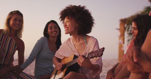 Group of friends sitting on sandy beach at sunset, one playing guitar, others smiling and enjoying music. Great for themes such as friendship, summer vacation, outdoor activities, relaxation, and music entertainment.