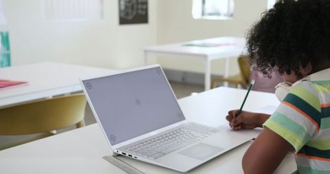 African american teen studying at desk with laptop and notebook, writing in classroom