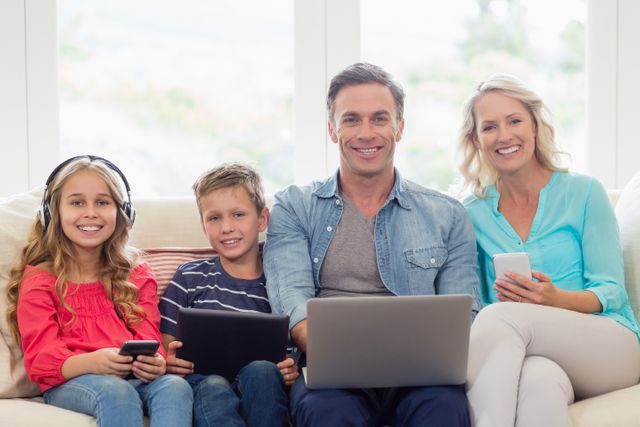 Portrait of smiling parents and kids sitting on sofa with laptop, mobile phone and digital tablet in living room