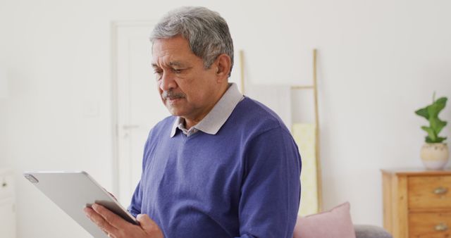 Image of senior biracial man using tablet standing in living room, smiling. Retirement, communication, inclusivity and senior lifestyle concept.