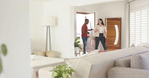 Couple entering sunlit home laughing while carrying bags and keys in modern living room