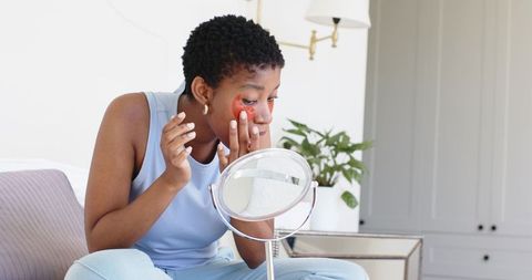 Applying under-eye hydrogel patch at vanity mirror, woman adjusting red eye mask in bright bedroom