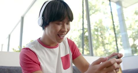 Asian student enjoying mobile game with headphones in sunny room